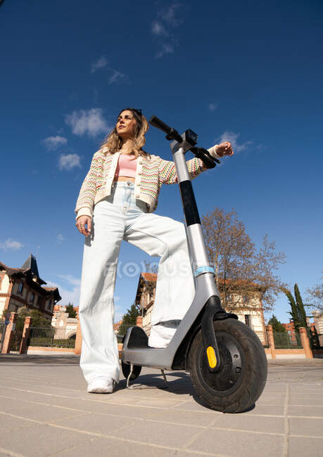 De baixo de jovem fêmea legal no desgaste da moda com scooter elétrico olhando para longe na passarela urbana — Fotografia de Stock