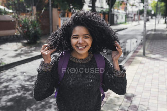 Crop view of delighted ethnic female student with Afro hairstyle and backpack standing on street on sunny day and looking at camera while touching her hair — Fotografia de Stock