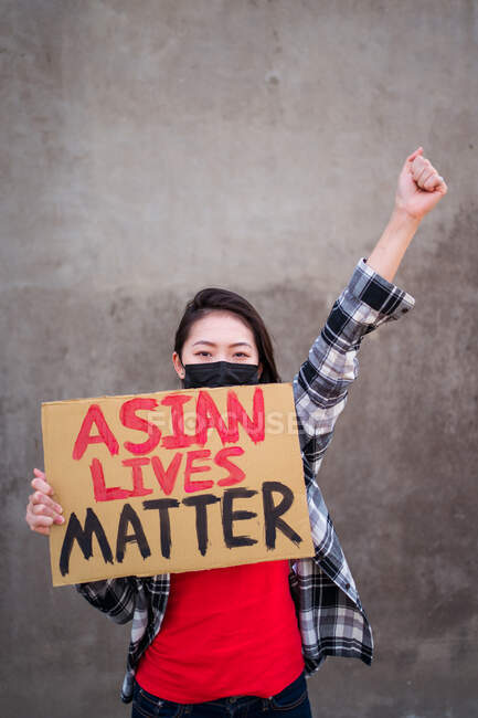 Donna etnica in maschera e con cartello di cartone con iscrizione Asian Lives Matter che protesta con braccio alzato in strada e guarda la macchina fotografica — Foto stock