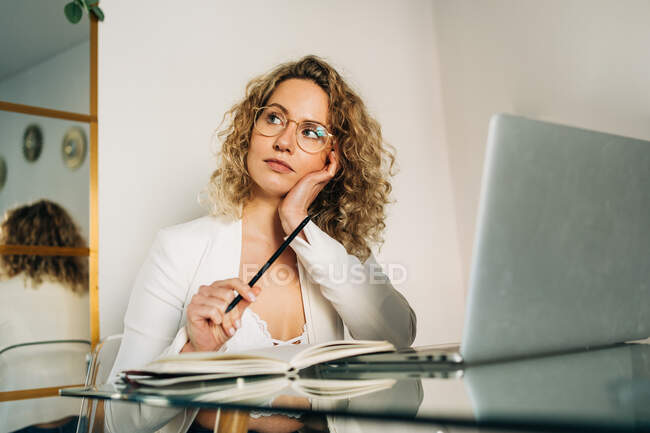 Задумчивая молодая женщина-фрилансер с кудрявыми светлыми волосами в повседневной одежде и очках делает заметки в планировщике и отворачивается, работая дистанционно с ноутбуком дома — стоковое фото