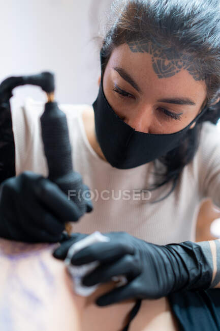 Female tattooer in cloth face mask with machine drawing tattoo on body of unrecognizable client in salon — Stock Photo