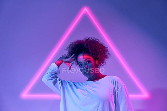 Retrato de confiante jovem afro-americana com cabelo encaracolado em pé olhando para a câmera em luzes de néon — Fotografia de Stock
