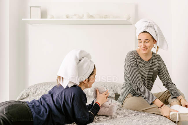 Счастливая молодая женщина завязывает шнурки, разговаривая с лучшей подругой в повседневной одежде и полотенцах на головах, расслабляясь на удобной кровати после купания дома — стоковое фото