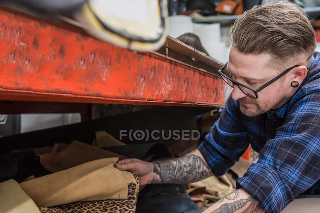 Bello tatuato maschio artigiano che sceglie i tessuti per la tappezzeria del sedile del motociclo mentre lavora in officina — Foto stock