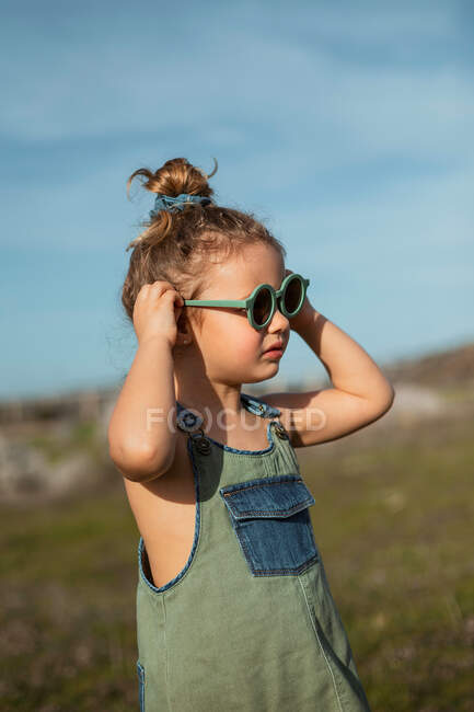Kleines Mädchen in Overalls und Sonnenbrille steht auf der Wiese und genießt den Sommer an einem sonnigen Tag im Grünen — Stockfoto