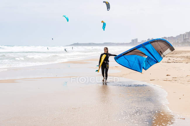 Desportista em roupa de mergulho com pipa inflável passeando na costa arenosa enquanto olha para longe contra o oceano tempestuoso — Fotografia de Stock