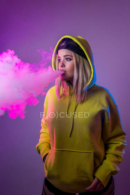 Femme confiante hipster en sweat à capuche fumer e cigarette en studio sur fond rose et détourner les yeux — Photo de stock