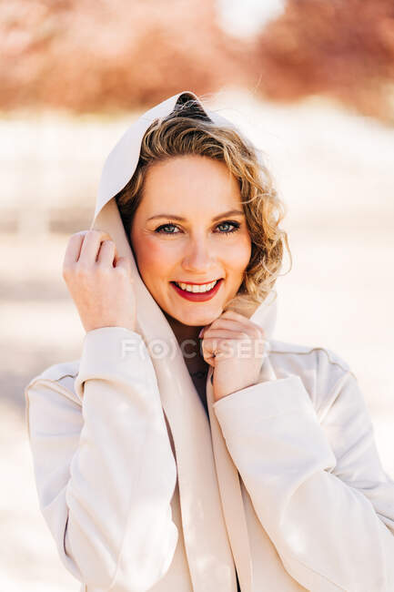 Femme heureuse en robe élégante et manteau marchant le long de l'allée entre les arbres et regardant la caméra — Photo de stock