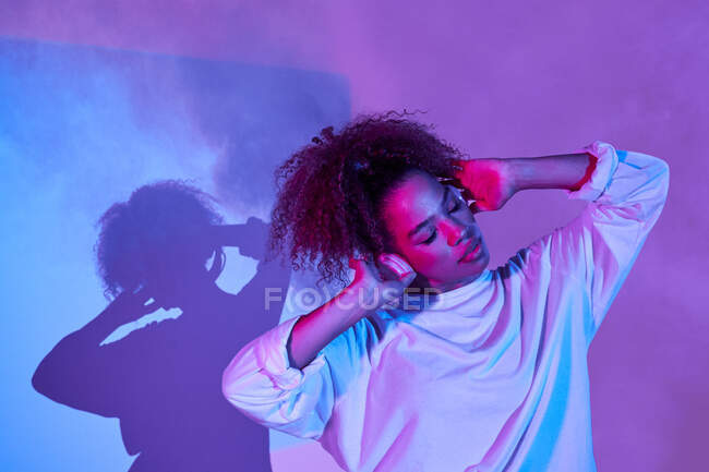 Nachdenkliche junge Afroamerikanerin in weißer Kleidung trägt Kopfhörer auf dem Hals, während sie mit geschlossenen Augen im dunklen Studio im Neonlicht steht — Stockfoto