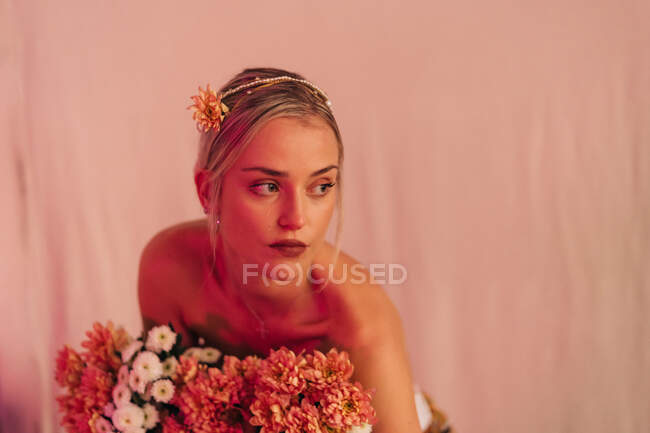 Charming romantic young bare shouldered female with bunch of fresh blooming flowers standing against beige background illuminated with neon light — Foto stock