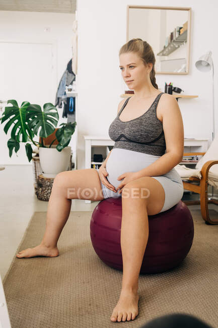 Junge werdende Frau streichelt Bauch, während sie auf Gymnastikball sitzt und im Wohnzimmer wegschaut — Stockfoto