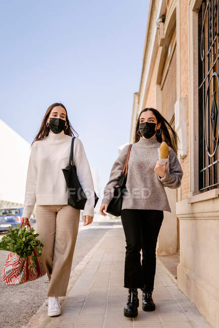 Stilvolle junge Freundinnen in lässiger Kleidung und Schutzmasken, die nach dem Einkauf im Lebensmittelgeschäft während der COVID-Pandemie auf der Straße der Stadt spazieren gehen — Stockfoto