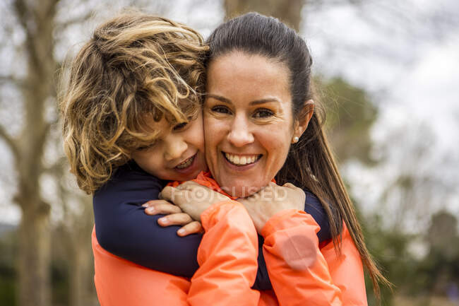 Charming boy embracing smiling mother while sitting in a wooden bench looking away in daylight - foto de stock