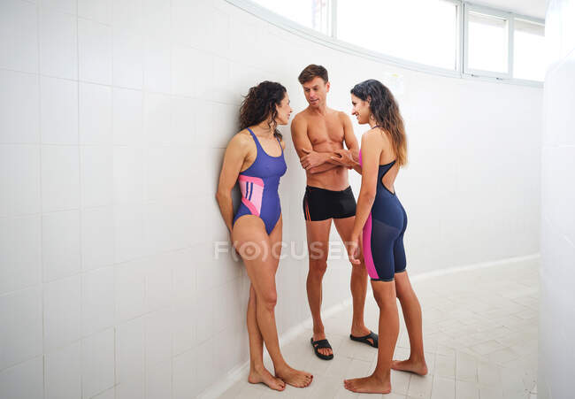 Sportsman with naked torso between female athletes in swimsuits speaking while standing on tiled floor in passage — Stock Photo