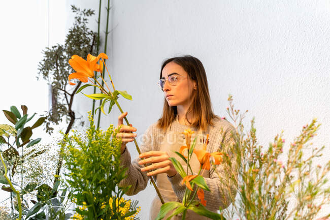Florista femenina joven concentrada en delantal y anteojos que arregla flores naranjas fragantes en jarrón mientras trabaja en una tienda de flores - foto de stock