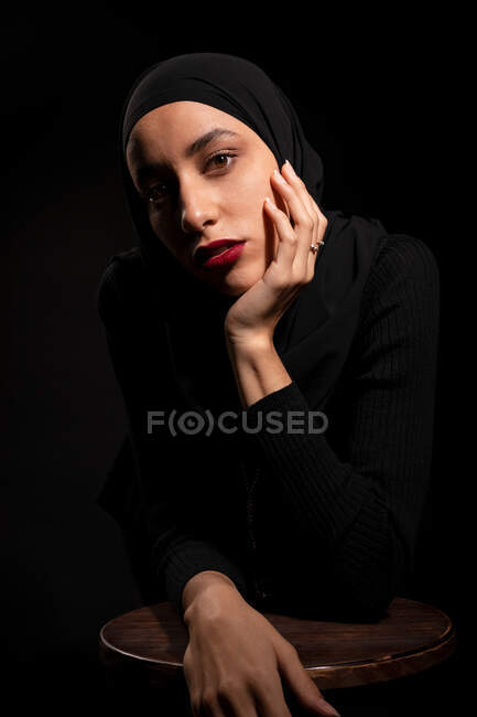 Attraente giovane donna islamica vestita di nero e hijab faccia toccante delicatamente appoggiata sulla sedia in studio nero guardando la fotocamera — Foto stock