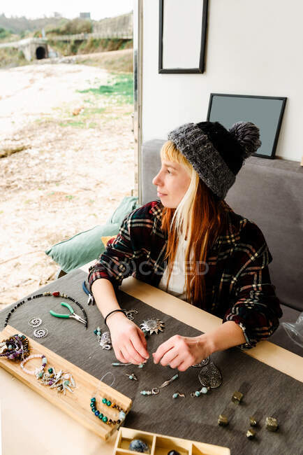Vista lateral del contenido viajero femenino creando accesorios hechos a mano mientras está sentado en la mesa de madera en el camión estacionado en la playa - foto de stock