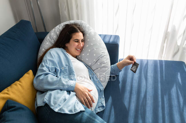 From above side view of pregnant female lying on sofa at home and looking at ultrasound scan photo of baby — Stock Photo