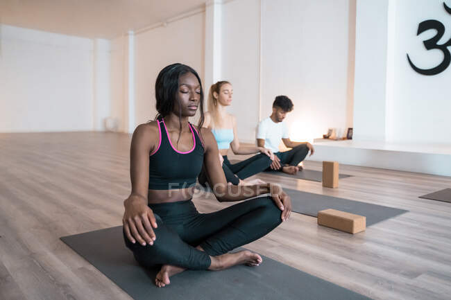 Donna afroamericana con gruppo di persone diverse sedute a Lotus posare con gli occhi chiusi e mediare durante la pratica dello yoga insieme durante la lezione in studio — Foto stock