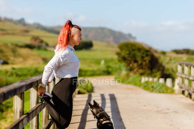 Vue latérale de l'athlète féminine souriante en vêtements de sport faisant de l'exercice sur un sentier clôturé tout en regardant loin par une journée ensoleillée — Photo de stock