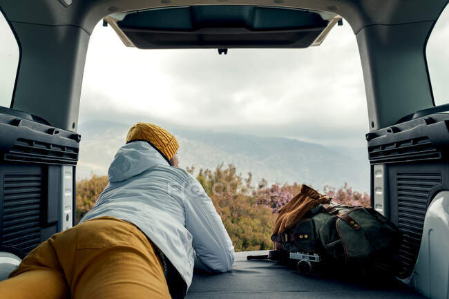 Задний вид на неузнаваемого мужчину-туриста в верхней одежде, лежащего на фургоне и любующегося живописным видом на высокогорье — стоковое фото