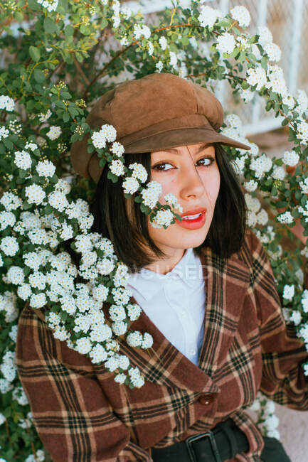 Молода ніжна етнічна жінка в картатому одязі з беретом, який торкається квітучої рослини, дивлячись на камеру на вулиці — стокове фото