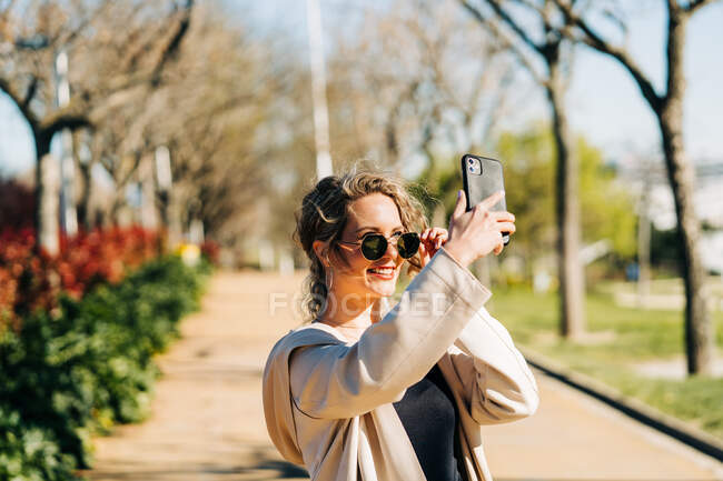 Весела жінка в модних сонцезахисних окулярах робить самостріл на смартфон, стоячи на стежці в саду в сонячний день — стокове фото