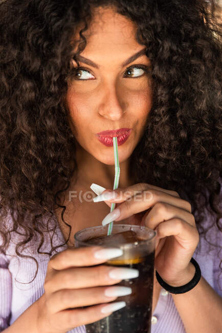 Crop feliz jovem fêmea com cabelos longos encaracolados bebendo bebida fria com palha e olhando para longe coquettishly — Fotografia de Stock