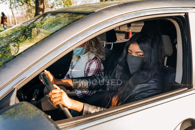 Conductor femenino negro en máscara sentado en automóvil moderno con amigo en la ciudad y mirando en el espejo lateral - foto de stock