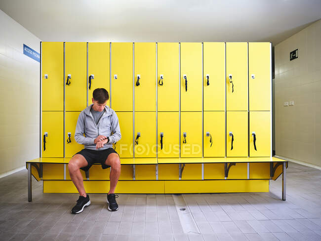 Взрослый спортсмен в спортивной одежде во время проверки времени, сидя на скамейке напротив желтых шкафчиков в светлом здании — стоковое фото