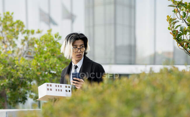 Giovane dirigente etnico maschile in occhiali con maquette casa e cellulare guardando altrove in città — Foto stock