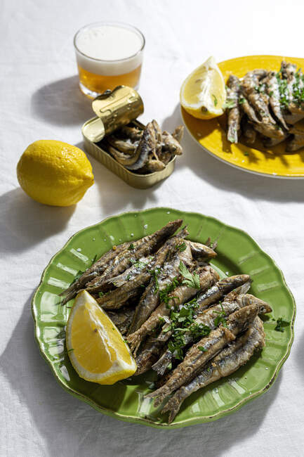 From above of delicious fried anchovies served on plates with lemon and placed on white table with glass of beer — Stock Photo