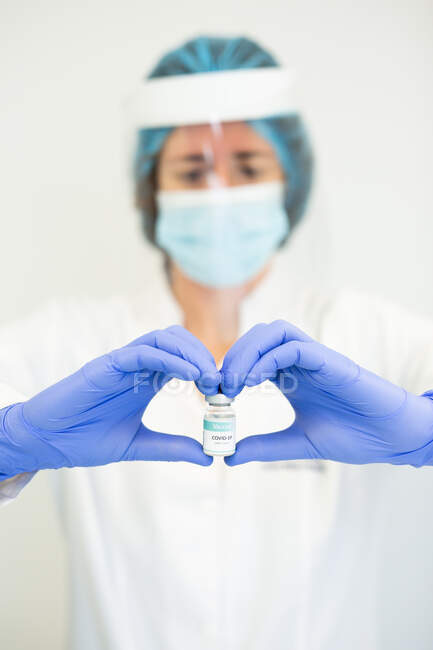 Doctora en guantes de látex y protector facial haciendo gesto en forma de corazón con las manos mientras sostiene líquido químico en un vial de vidrio en la clínica durante el brote de coronavirus - foto de stock