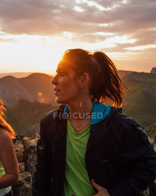 Morena em jaqueta esportiva em pé na parte de trás iluminado e olhando para longe contra cume da montanha enquanto explora a natureza com amigo — Fotografia de Stock
