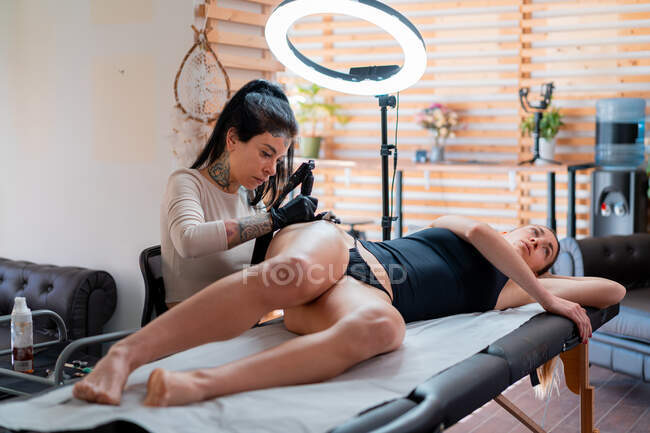 Tatoueuse dans des gants avec machine professionnelle appliquant le tatouage sur le corps de la femme dans le salon — Photo de stock