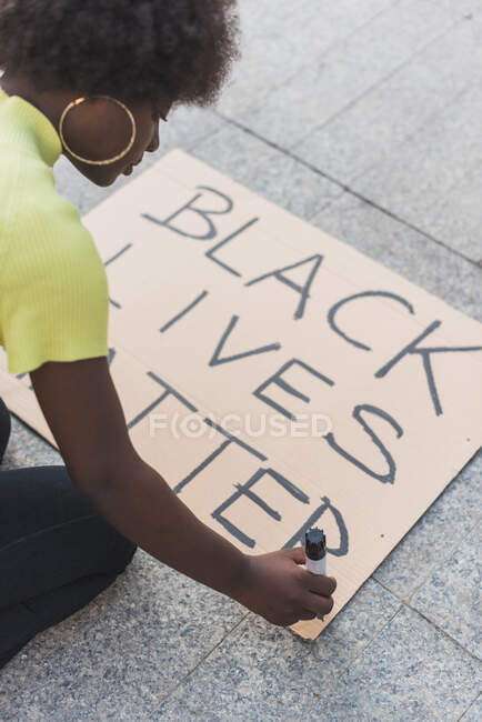 High angle side view of African American female activist writing Black Lives Matter and making placard for protest against racism in city — Photo de stock