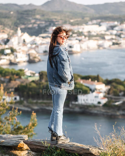 Back view full body content female standing on hilly seafront and admiring sea and coastal town views on windy day — Stock Photo