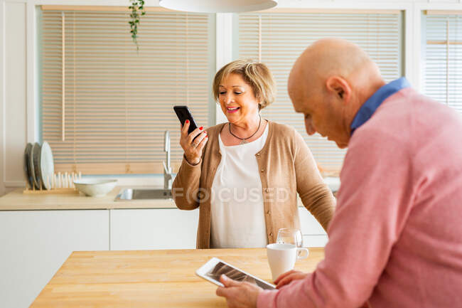 Содержание пары средних лет просмотр планшета и смартфона утром, стоя за столом на кухне — стоковое фото