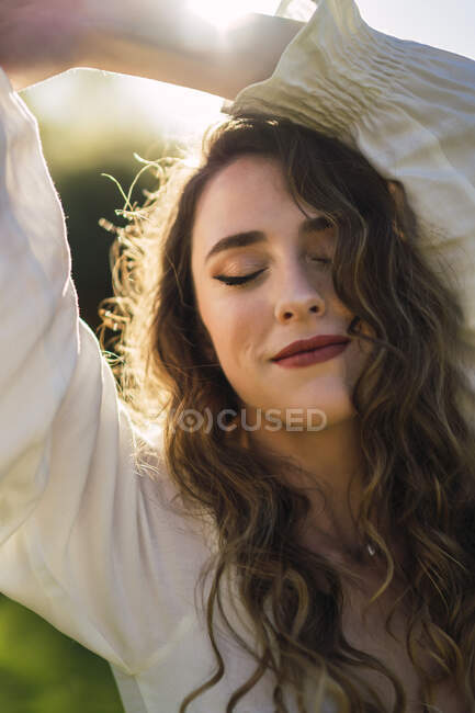 Beautiful content female with closed eyes and long curly hair raising arms gracefully with smile while spending sunny day in verdant nature — Stock Photo