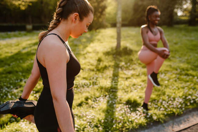 Young multiracial female athletes in sportswear stretching arm and legs on asphalt footpath in town on sunny day — Stock Photo