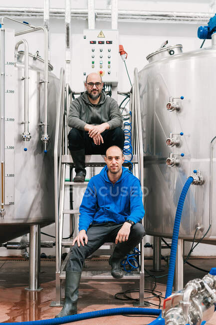 Zufriedene männliche Mitarbeiter blicken in die Kamera, während sie in Brauerei auf Treppen zwischen Metalltanks sitzen — Stockfoto