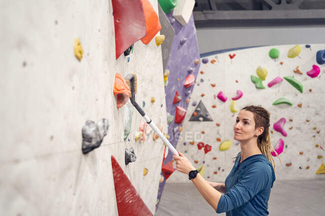 Lächelnde Alpinistin reinigt Felsen an künstlicher Wand mit Pinsel in modernem Boulderzentrum — Stockfoto