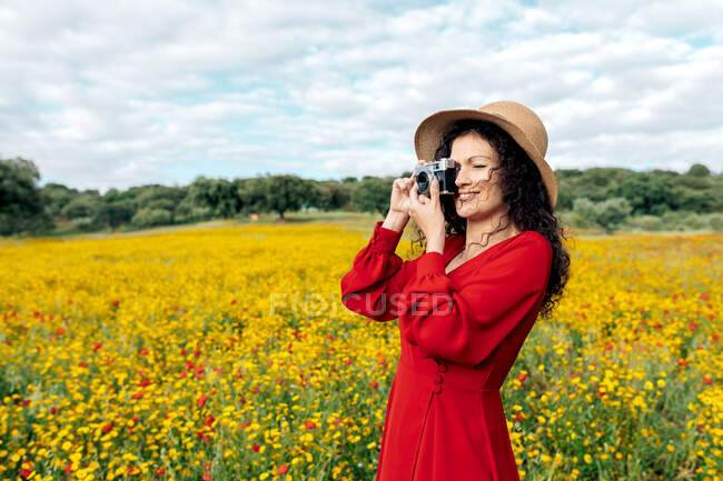 Усміхнена жінка в капелюсі фотографується на старовинній камері на лузі під хмарним небом — стокове фото