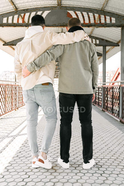 Back view of unrecognizable young gay couple in stylish clothes hugging each other while standing on bridge on sunny day — Foto stock