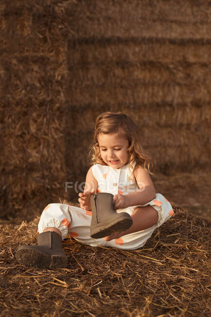 Adorable curious child in overalls sitting on haystack and looking at boots in evening in countryside — Stock Photo