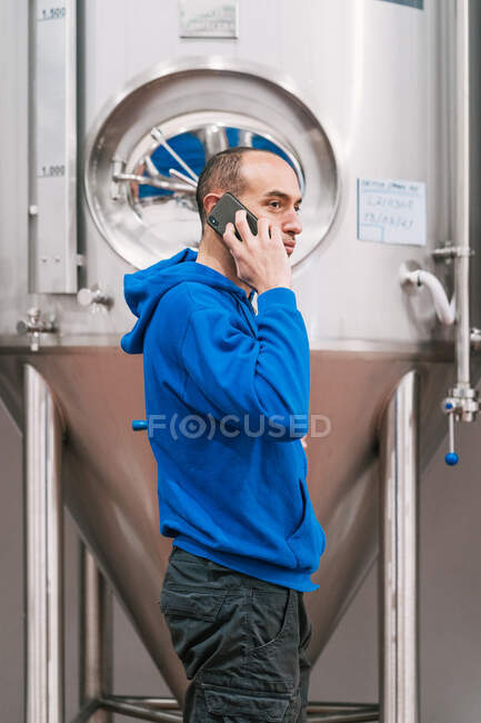 Side view of male entrepreneur talking on cellphone against stainless steel vessels on wet floor in beer factory — Stock Photo