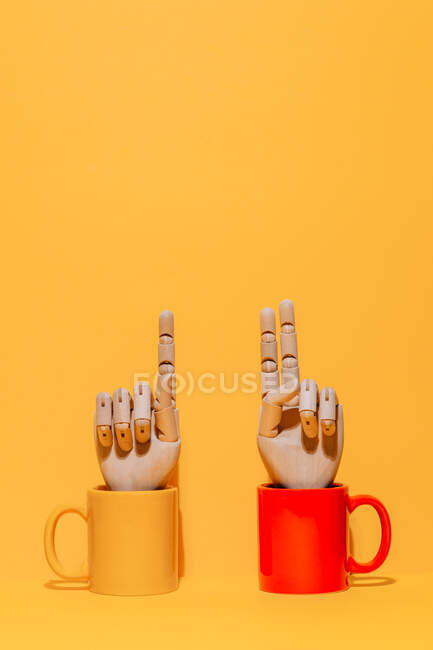 Creative ornamental wooden hand with index and middle finger together gesture inside colorful mug on yellow and red background in studio — Stock Photo