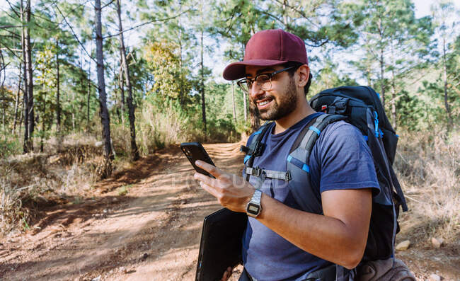 Feliz joven caminante masculino étnico en ropa casual y gorra con mochila y computadora portátil que navega por el teléfono móvil en el exuberante bosque de verano - foto de stock