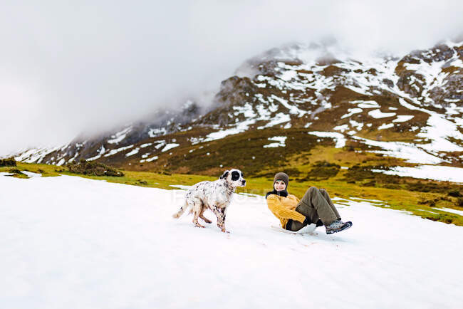 Happy woman sliding down slope covered with snow while happy English Setter dog running aside in mountains — Stock Photo