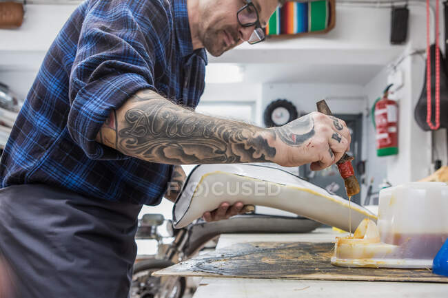 Seitenansicht tätowierter Handwerker, der beim Polstern an der Werkbank Kleber auf Motorradsitz aufträgt — Stockfoto